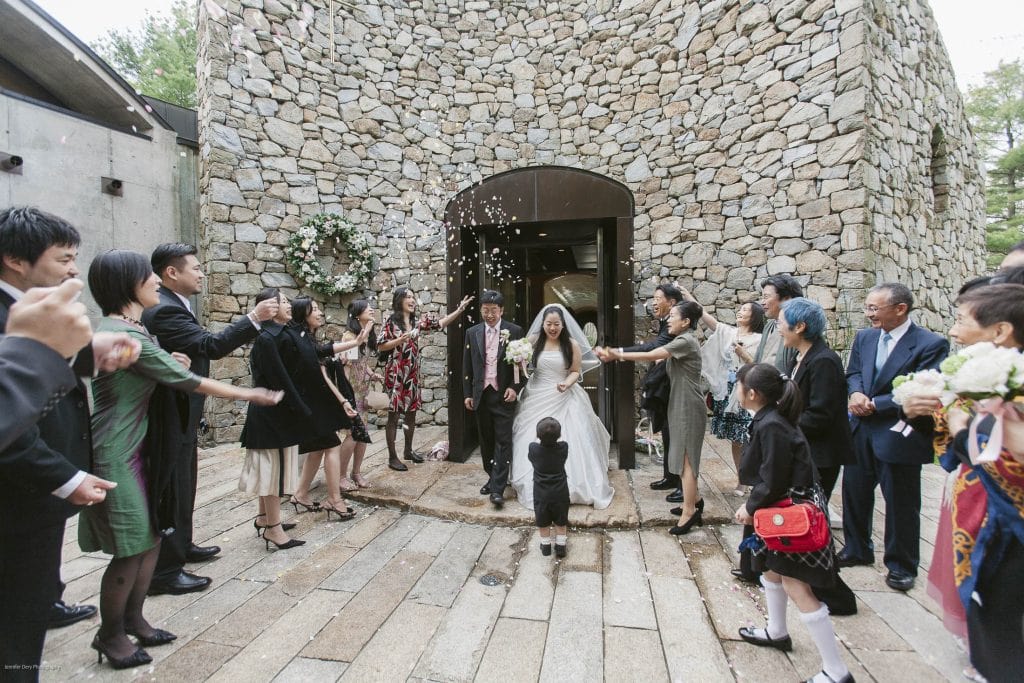 A bride and groom exit a stone building as guests on both sides throw flower petals and celebrate; a child stands in front of the couple, and everyone is dressed formally.