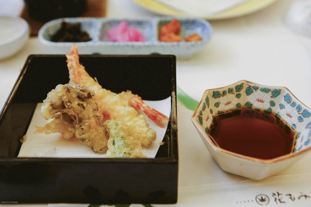 A black bento box with assorted tempura, including shrimp and vegetables, sits next to a small bowl of dipping sauce. In the background, a tray holds small portions of pickled vegetables.