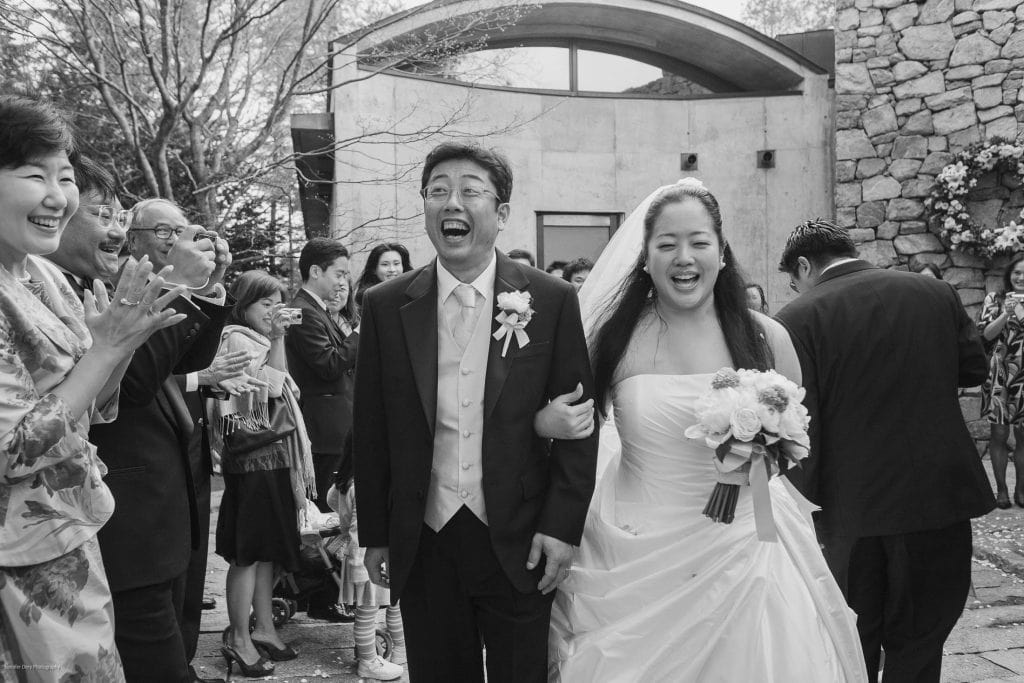 A joyful bride and groom walk arm in arm down an outdoor aisle, smiling and laughing as guests applaud and celebrate their wedding. The scene is captured in black and white.