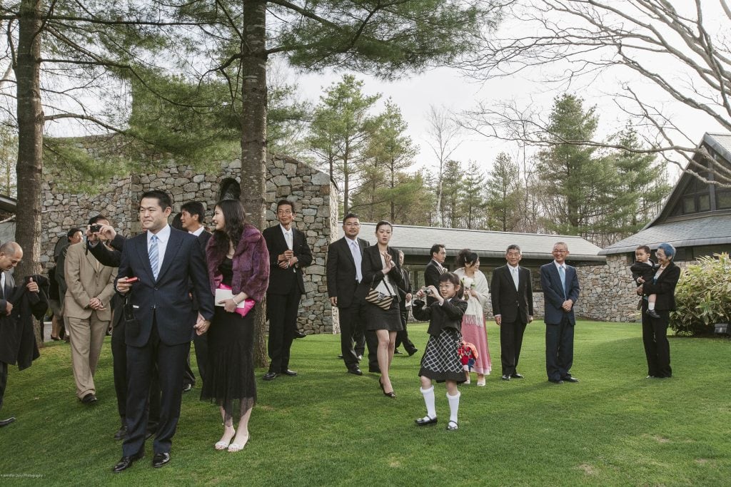 A group of people dressed formally gathers on a lawn, some smiling and talking, while a young girl in a black dress stands in front clapping. Trees and stone buildings are visible in the background.