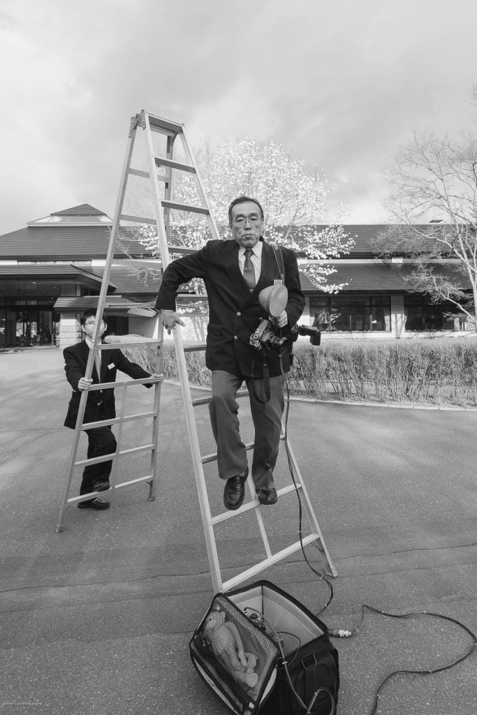 A man in a suit stands on a stepladder holding a camera and equipment, with another person holding the ladder. The scene is outdoors near a building with trees and a suitcase on the ground. The image is in black and white.