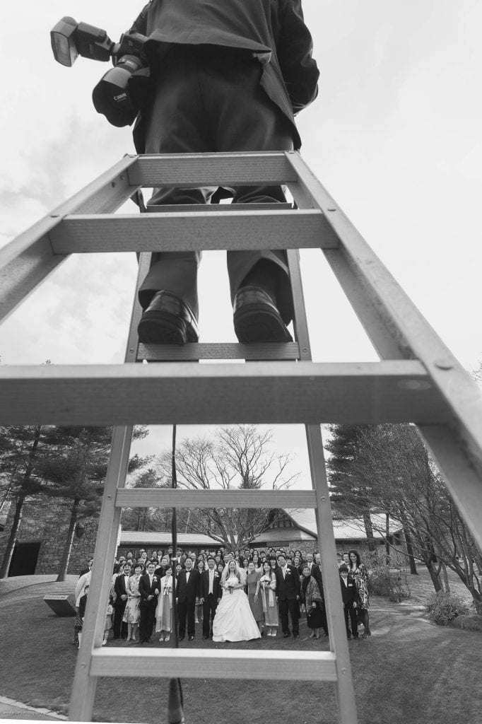 A photographer stands on a tall ladder, capturing a group wedding photo below. The image is shot from beneath the ladder, framing the bride, groom, and guests gathered outdoors.