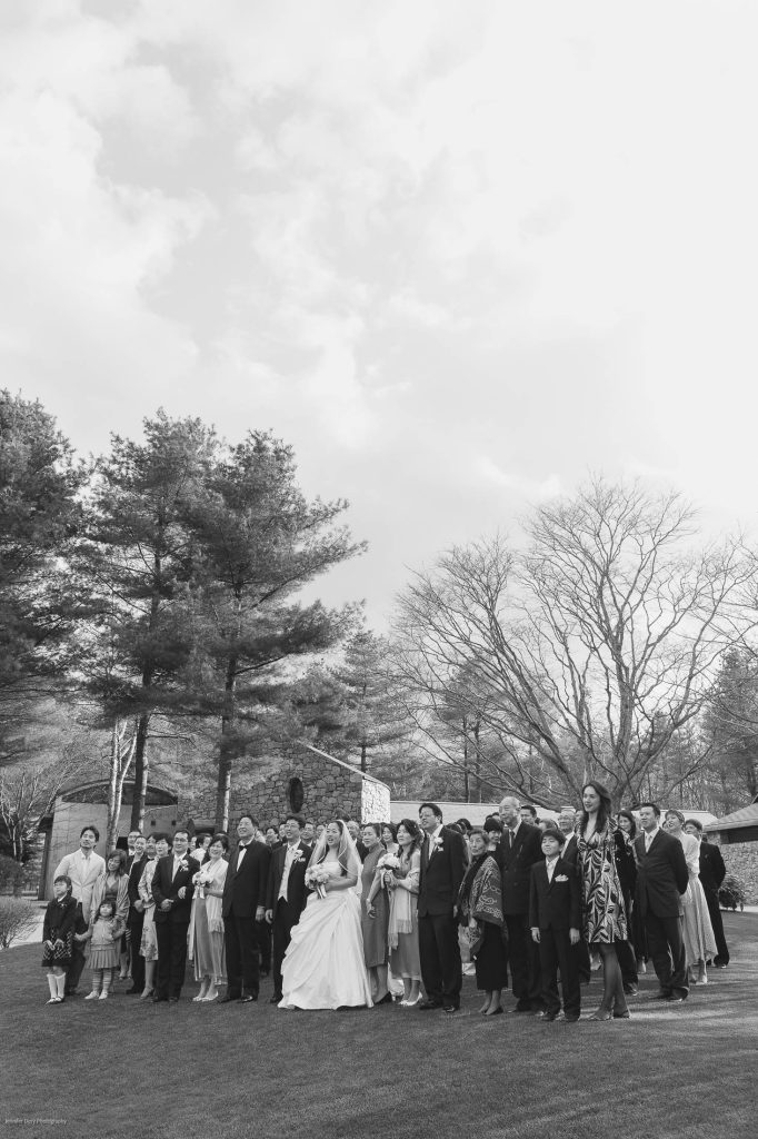 A large group of people, including a bride in a white dress, stand on a lawn outdoors, looking upward. Tall trees and a cloudy sky make up the background. The image is in black and white.