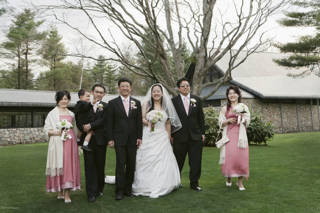 Six people, including a bride and groom, pose for a wedding photo outside on green grass, with trees and a stone building in the background. The group is smiling, dressed in formal attire, and holding bouquets.
