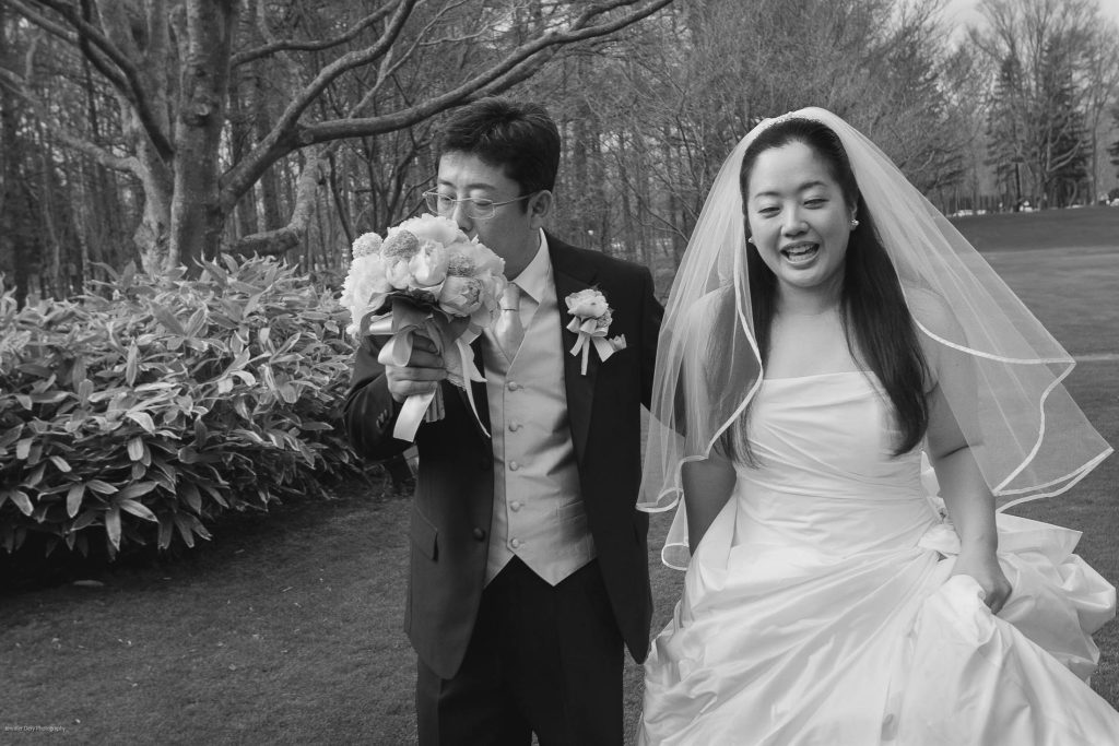 A bride in a white dress and veil walks on grass, smiling, while a groom in a suit and boutonniere sniffs a bouquet of flowers beside her. Trees and shrubs are in the background.