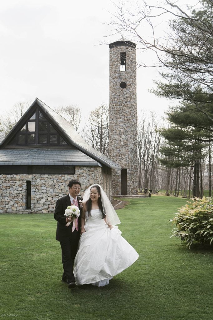 A bride and groom walk arm in arm on green grass, smiling and dressed in wedding attire, with a bouquet in hand. Behind them is a stone building and a tall stone tower surrounded by trees.