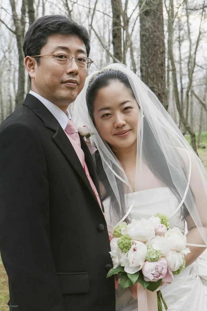 A bride and groom pose outdoors among trees. The bride wears a white dress, holds a bouquet of flowers, and has a veil over her head. The groom is in a black suit with a pink tie and boutonniere. Both look at the camera.