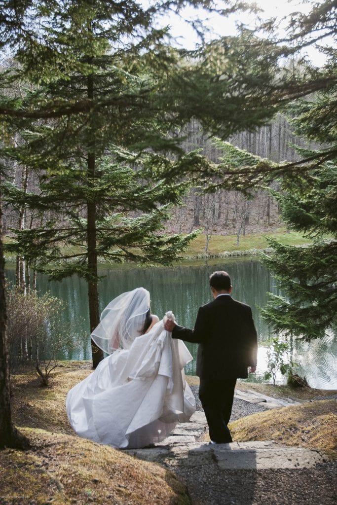 A bride and groom walk hand in hand down a stone path toward a serene lake, surrounded by tall pine trees and soft sunlight filtering through the branches.