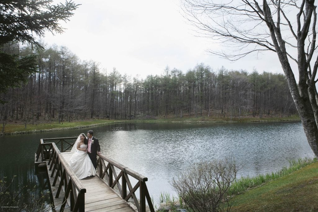 A bride and groom stand together on a wooden bridge over a small lake, surrounded by trees and greenery under a cloudy sky. The scene is peaceful and romantic.