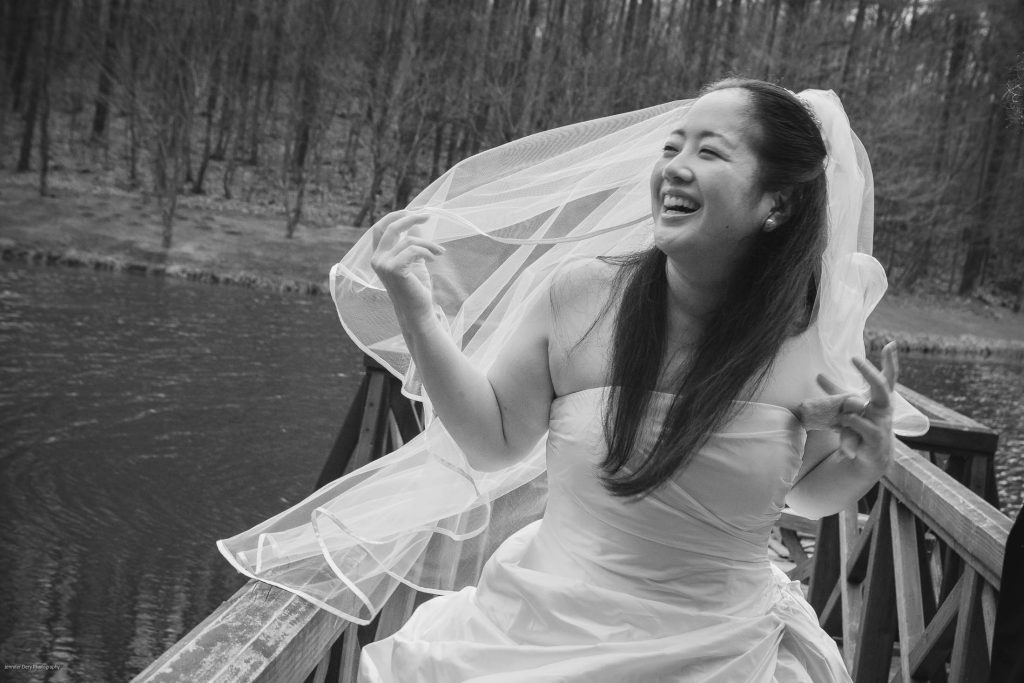 A joyful bride in a strapless wedding dress and veil laughs while standing on a wooden bridge over a pond, with bare trees in the background. The image is in black and white.