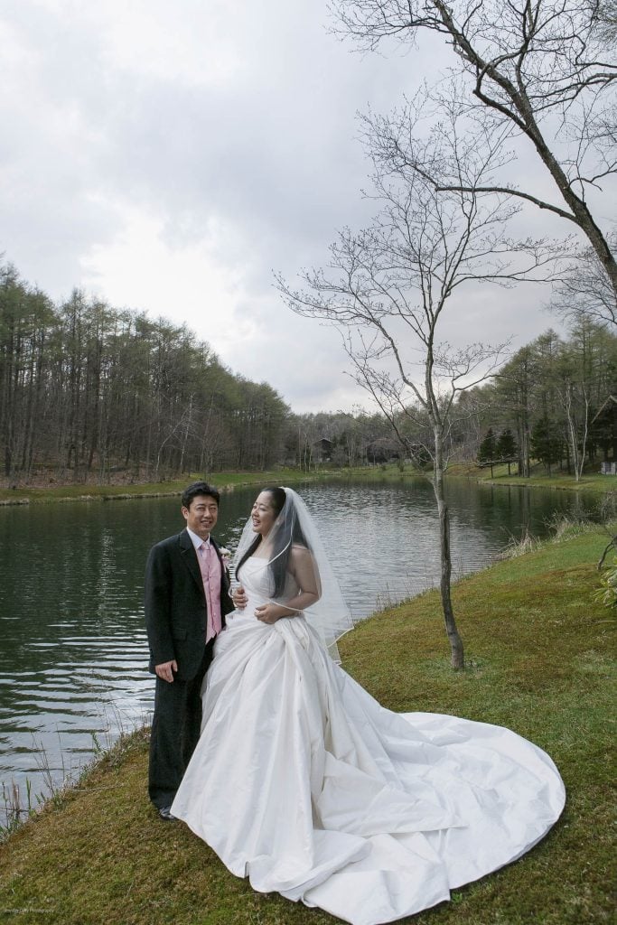 A bride in a long white dress and veil stands smiling beside a groom in a black suit by a peaceful lake, surrounded by trees and grass under a cloudy sky.