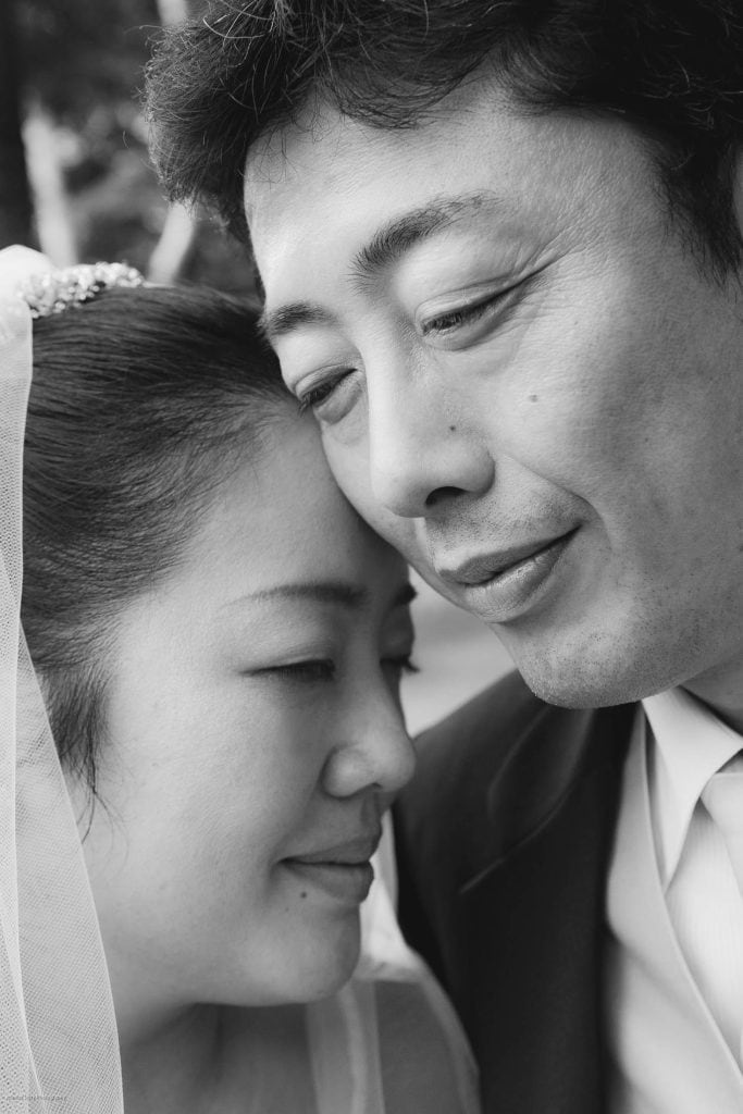 A close-up black-and-white photo of a bride and groom, their faces gently touching, both smiling softly with eyes closed, conveying a sense of love and intimacy on their wedding day.