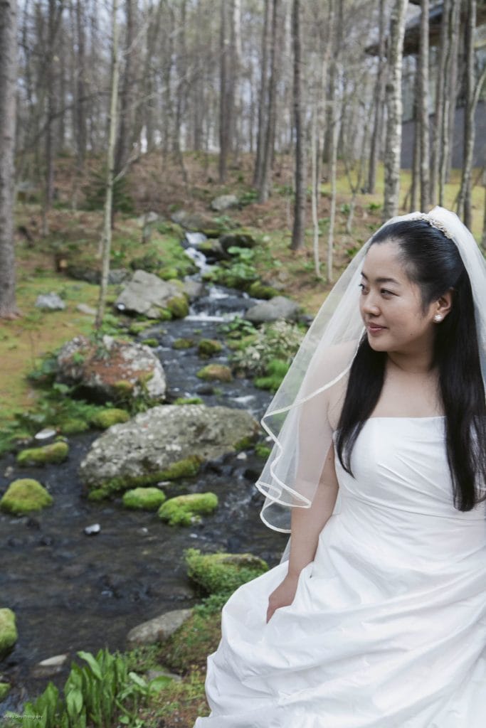 A bride in a white wedding dress and veil sits beside a small stream in a forest, surrounded by moss-covered rocks and trees. She is looking to the side and smiling gently.