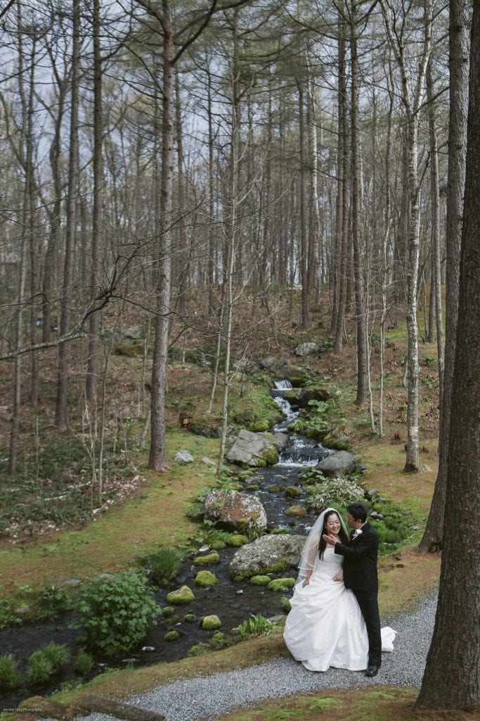 A bride and groom stand smiling and embracing on a gravel path in a forest, with a small stream and rocks flowing behind them among tall, leafless trees.