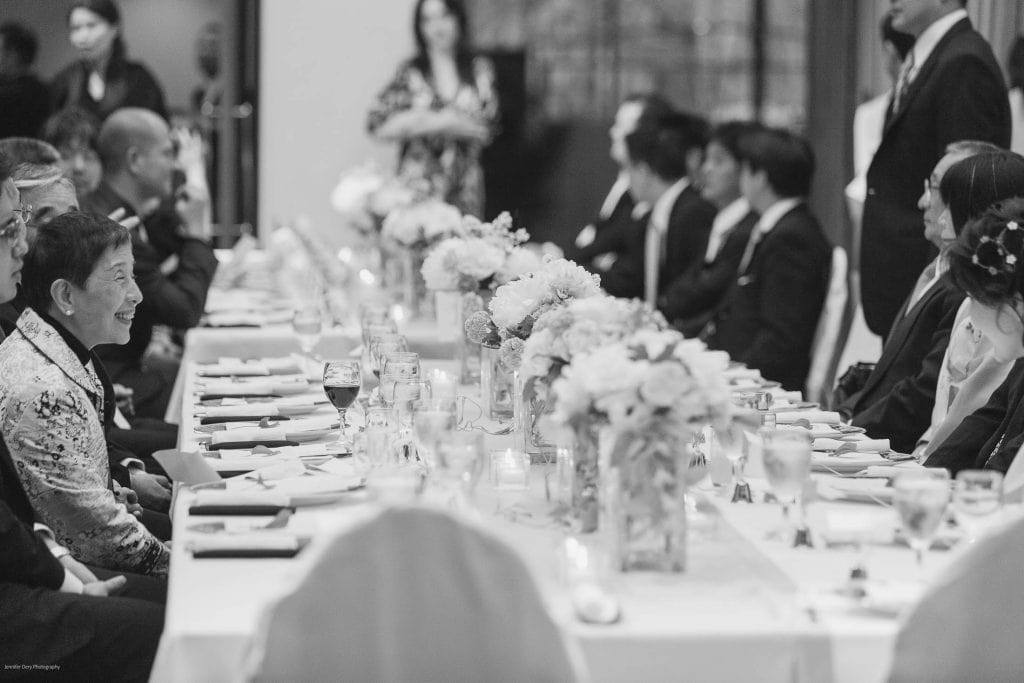 A black and white photo shows people dressed formally, seated on both sides of a long table decorated with flowers and glasses, as they smile and engage in conversation at a gathering or celebration.