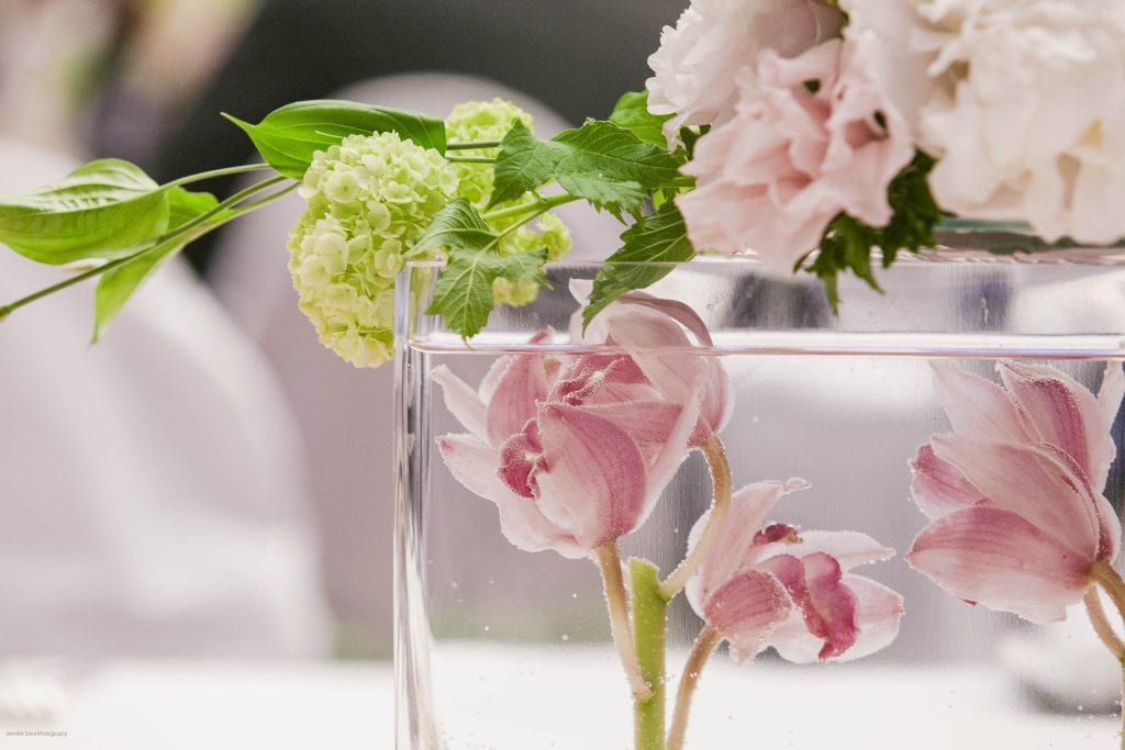 A close-up of pale pink flowers and green leaves arranged in a clear glass vase, with some flowers submerged in water and others resting on the rim, against a softly blurred background.