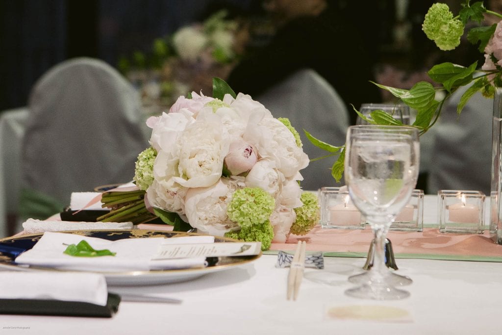 A bouquet of white and pale pink flowers with greenery rests on an elegantly set table with a glass of water, plates, chopsticks, candles, and a soft pink table runner.