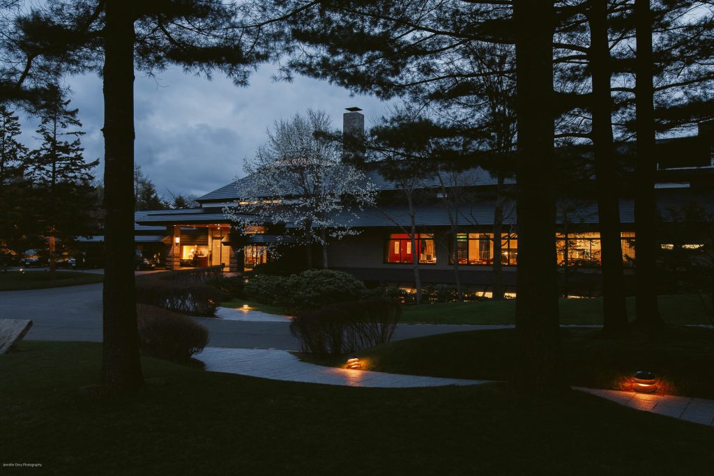 A modern house at dusk with warm lights glowing from the windows and pathway lights illuminating the curved walkway, surrounded by trees and landscaped bushes.