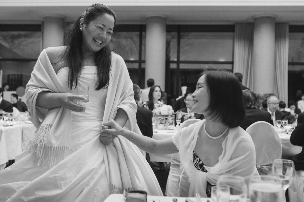 A bride in a white dress smiles and holds hands with a seated woman at a formal indoor event. Guests sit at tables in the background, and both women appear joyful and engaged in conversation.