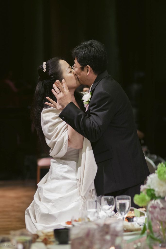 A bride and groom share a kiss at their wedding reception, dressed in formal attire. They stand beside a decorated table with flowers, glasses, and plates visible in the foreground.