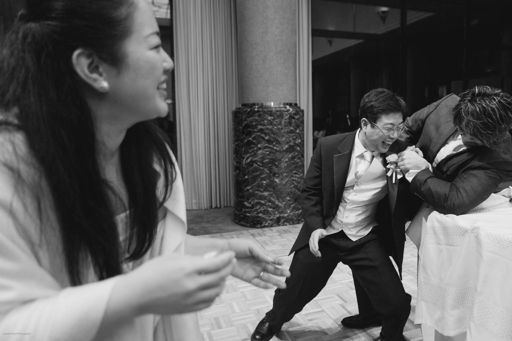 A woman laughs in the foreground while two men in suits playfully interact, with one man holding the other’s lapel as they both smile, in a lively indoor setting.