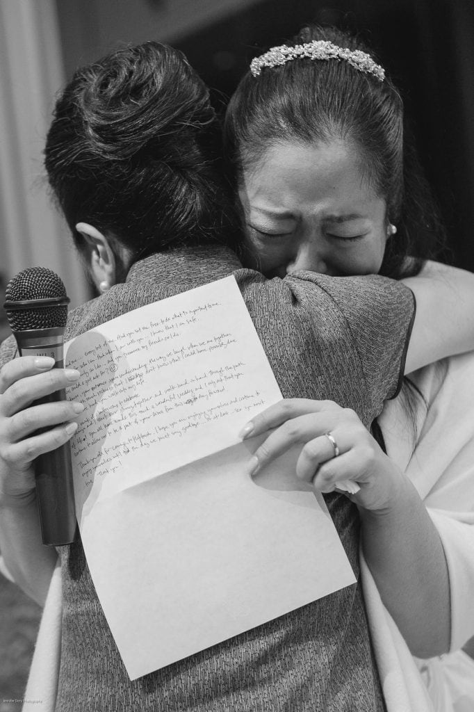 Two women embrace tightly, one holding a handwritten letter and a microphone. The woman facing the camera is visibly emotional, eyes closed and tearful, while the other’s face is turned away. The scene is captured in black and white.