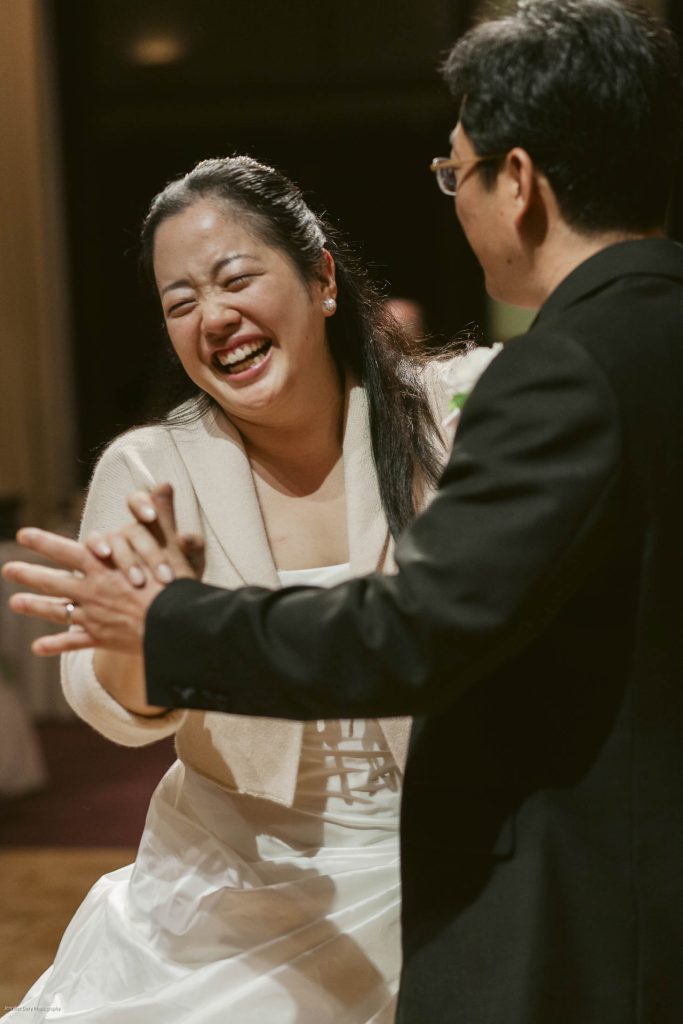 A joyful woman in a white dress and light jacket laughs while dancing with a man in a dark suit. They are holding hands and appear to be enjoying a special moment together at an event or celebration.