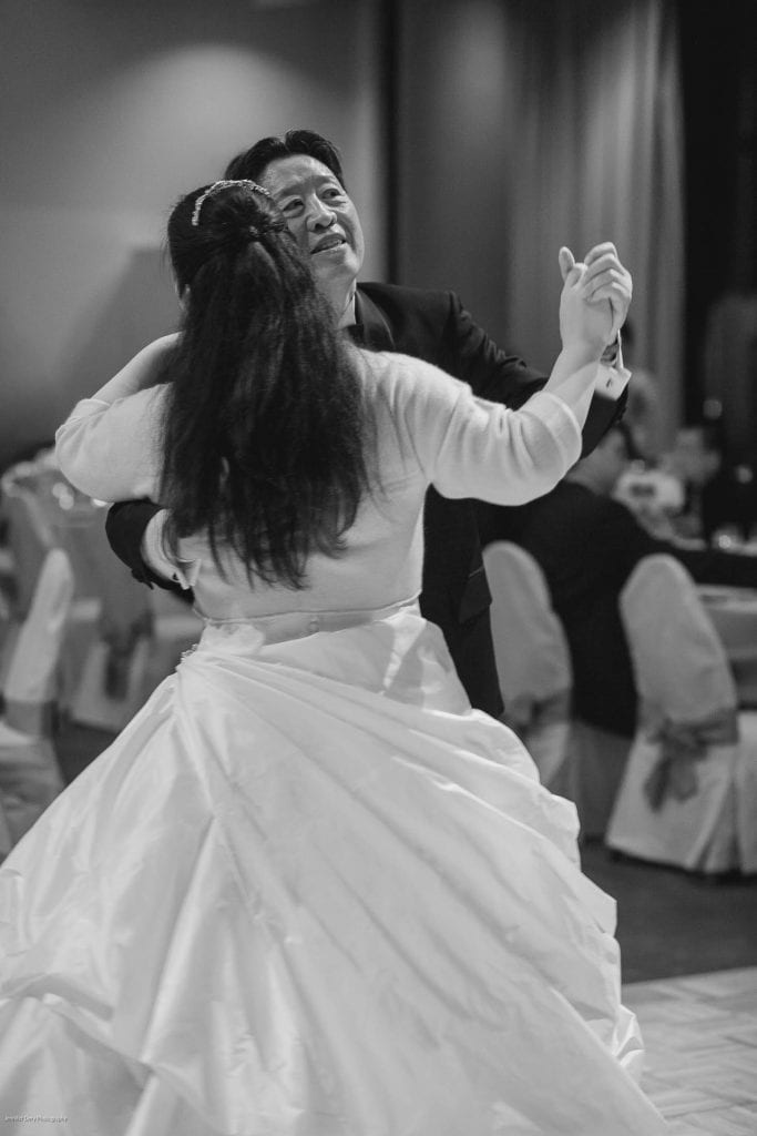 A bride and groom dance together at their wedding reception. The groom is smiling while holding the bride, who is wearing a flowing gown. The scene is in black and white, with guests seated in the background.