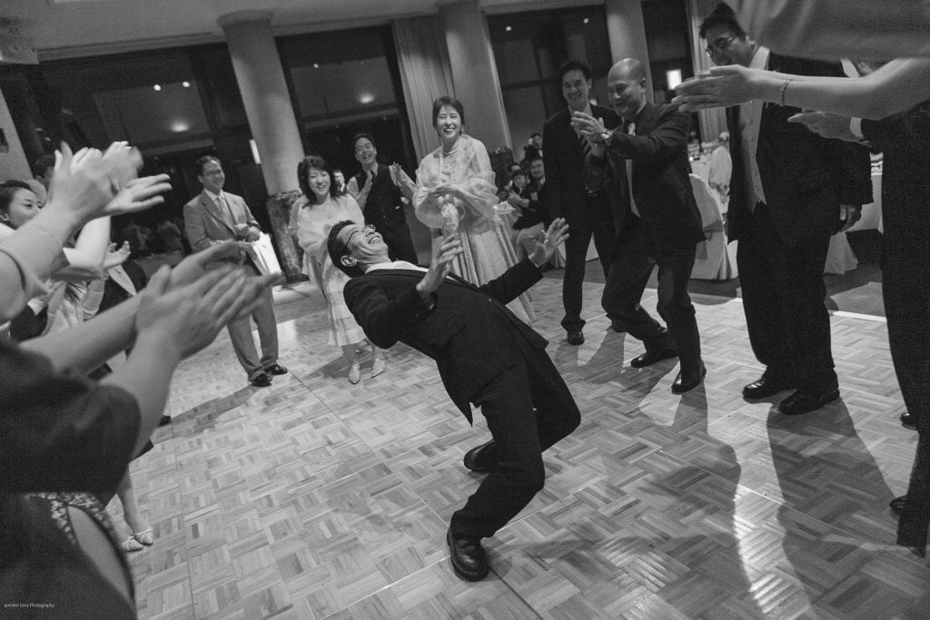 A man in a suit leans back to perform the limbo at an indoor party, while guests around him cheer, clap, and smile. The scene is lively and joyful, with people dressed formally.