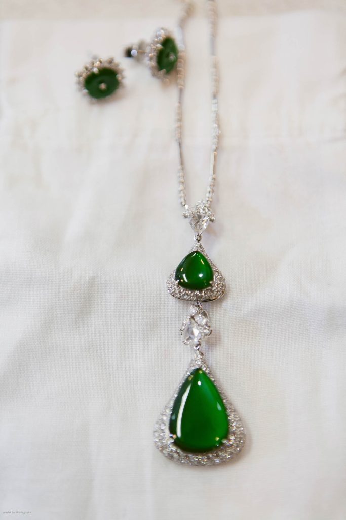 A silver necklace with two green teardrop-shaped gemstones surrounded by small clear stones, displayed on white fabric, with matching green gemstone earrings in the background.
