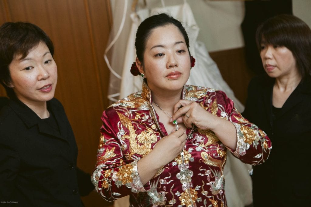 A woman in a traditional red and gold embroidered dress fastens a necklace, with two women in black standing beside her. A white dress hangs in the background.