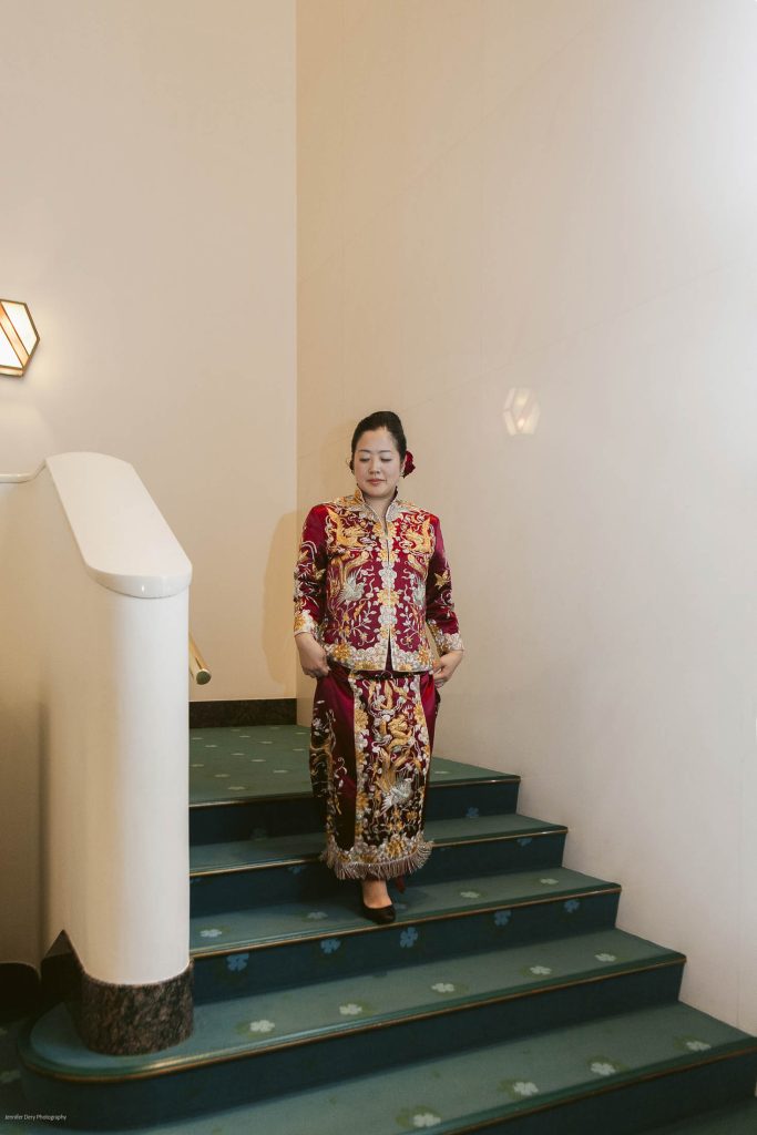 A woman wearing a traditional, ornate red and gold embroidered outfit stands barefoot on a staircase with green carpet, holding the sides of her garment. The setting appears to be indoors with light-colored walls.
