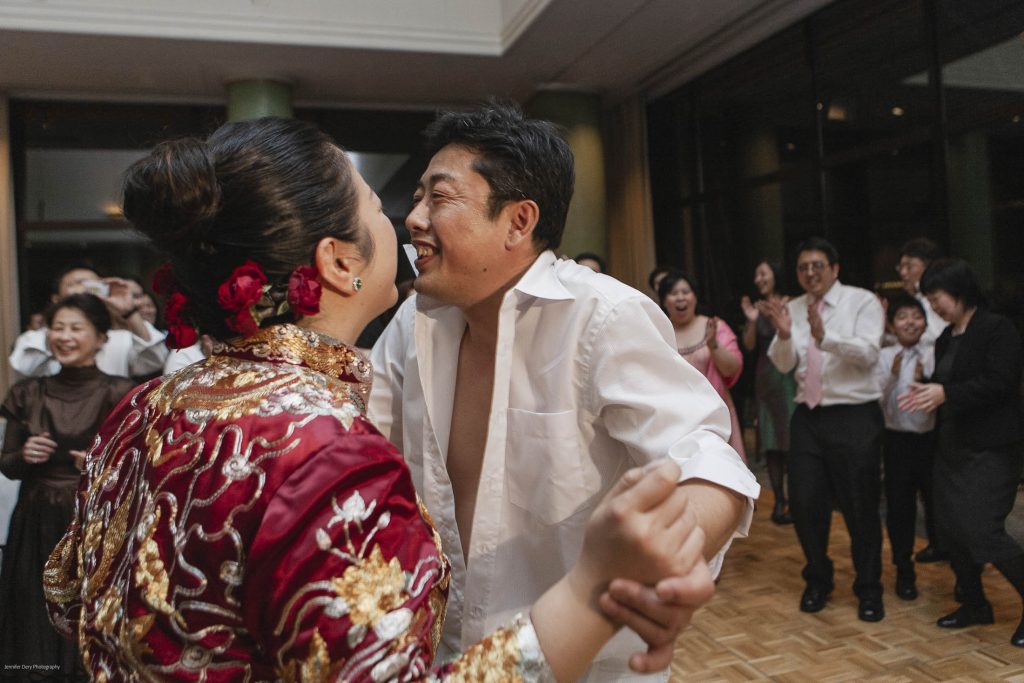 A couple dances joyfully at a celebration, with the woman in a red floral dress and the man in an open white shirt. Guests in the background cheer and clap, creating a lively and festive atmosphere.