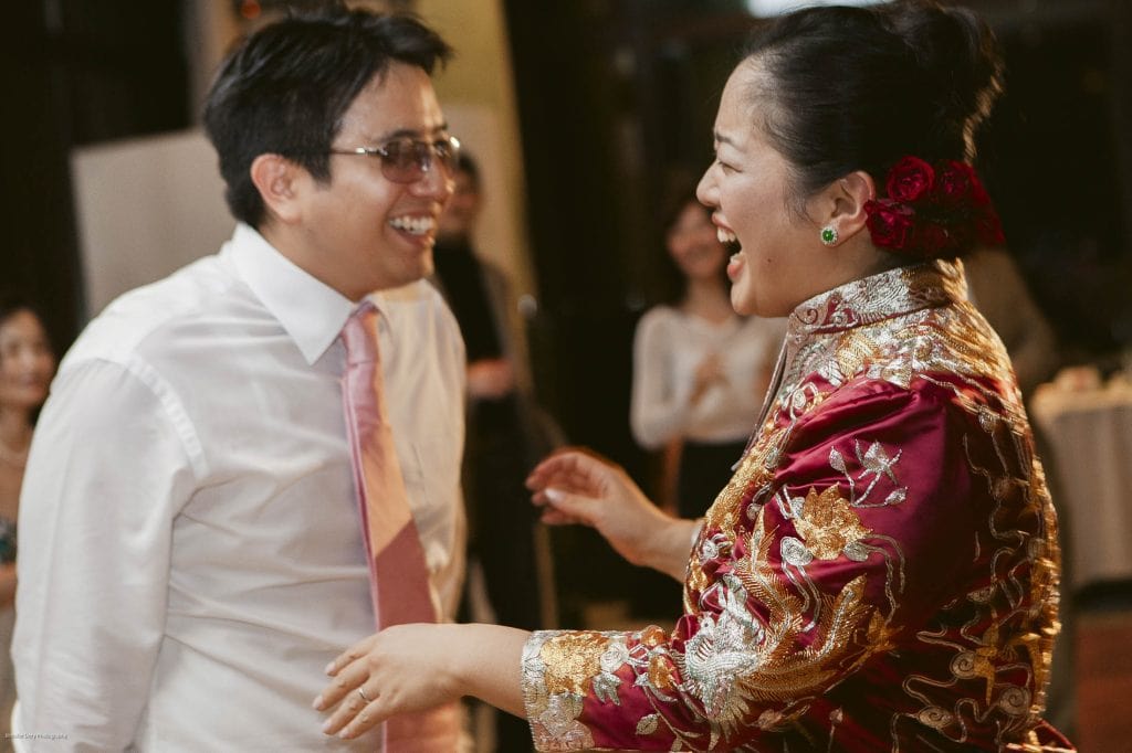 A man in a white shirt and pink tie laughs with a woman in a red and gold traditional dress, adorned with red flowers in her hair, both smiling and enjoying a joyful moment at a celebration.