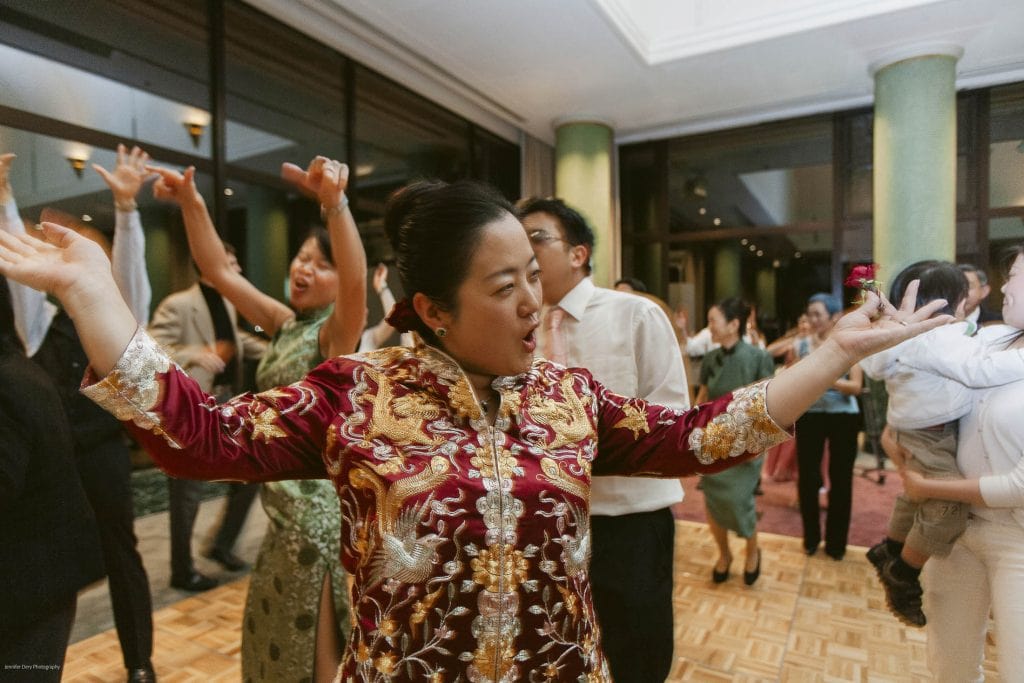 A woman in a red and gold embroidered dress dances with her arms raised at a lively indoor celebration, surrounded by people also dancing and enjoying the event.