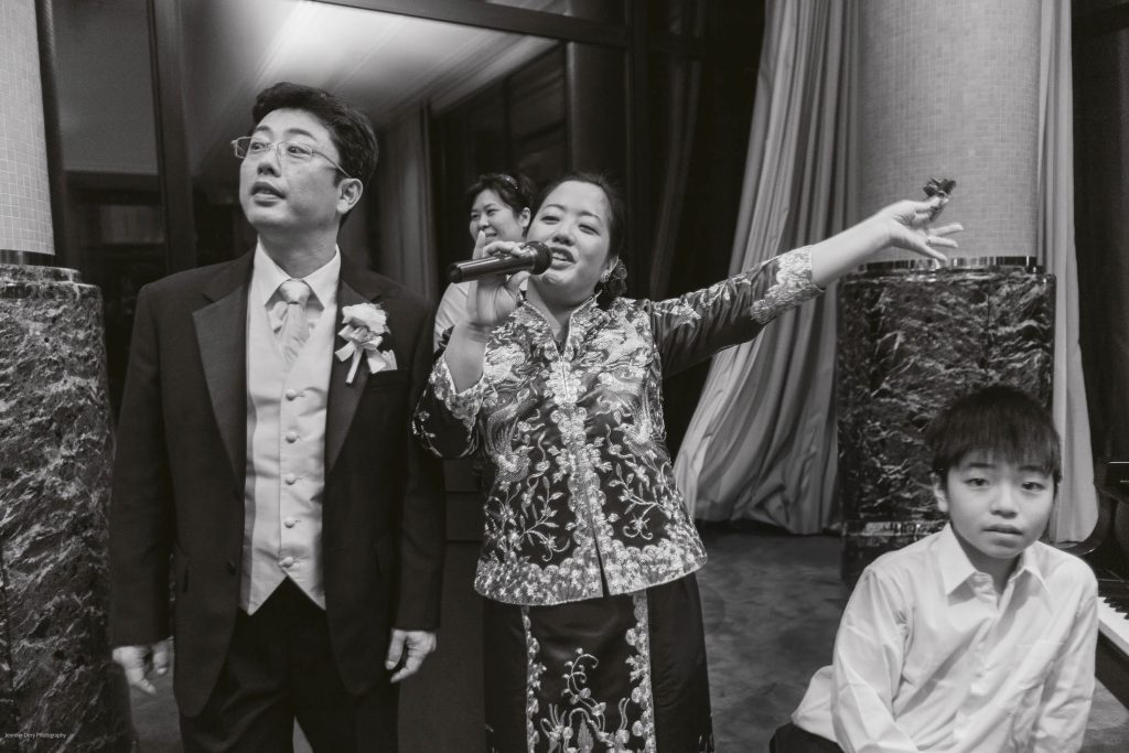 A woman in traditional attire sings into a microphone with her arm raised, standing beside a man in a suit and a boy in a white shirt; another woman stands in the background. All appear to be at a formal event.
