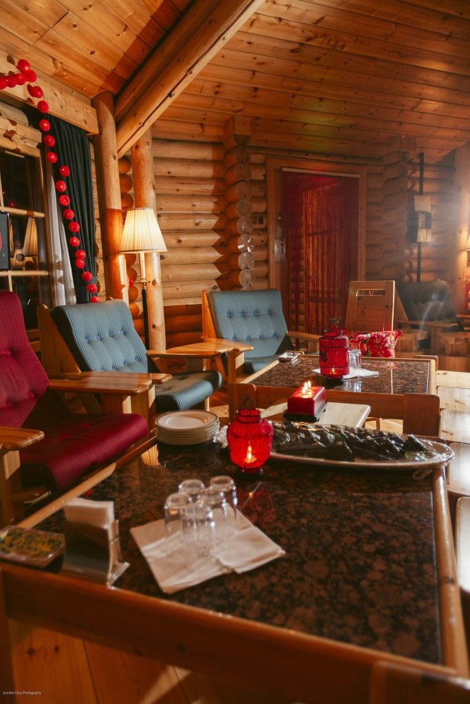 Cozy wooden cabin interior with comfortable chairs, a lamp, red decorations, and tables set with glasses, plates, and red candles, creating a warm and inviting atmosphere.