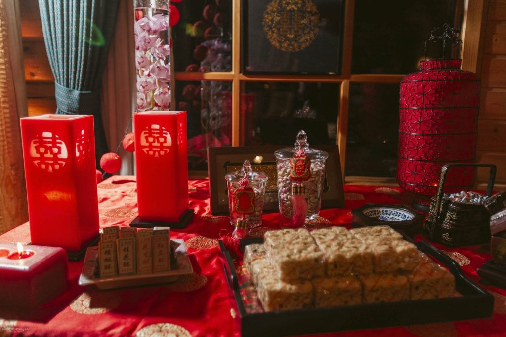 A festive table decorated with red candles, ornate jars, traditional sweets, and Chinese wedding symbols, set on a red cloth with gold accents and lit by warm indoor lighting.