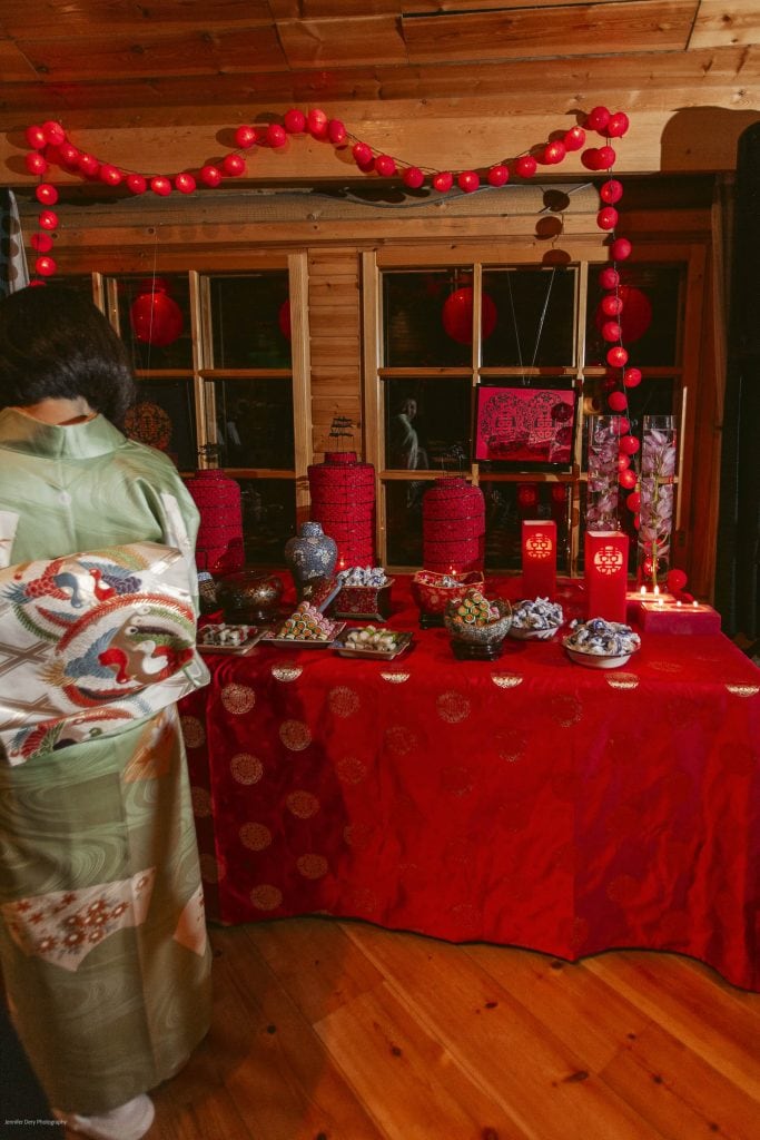 A person in a green and white kimono stands by a red-decorated table with vases, jars, and lanterns. Red lanterns and garlands hang above, creating a festive atmosphere in a wooden room.