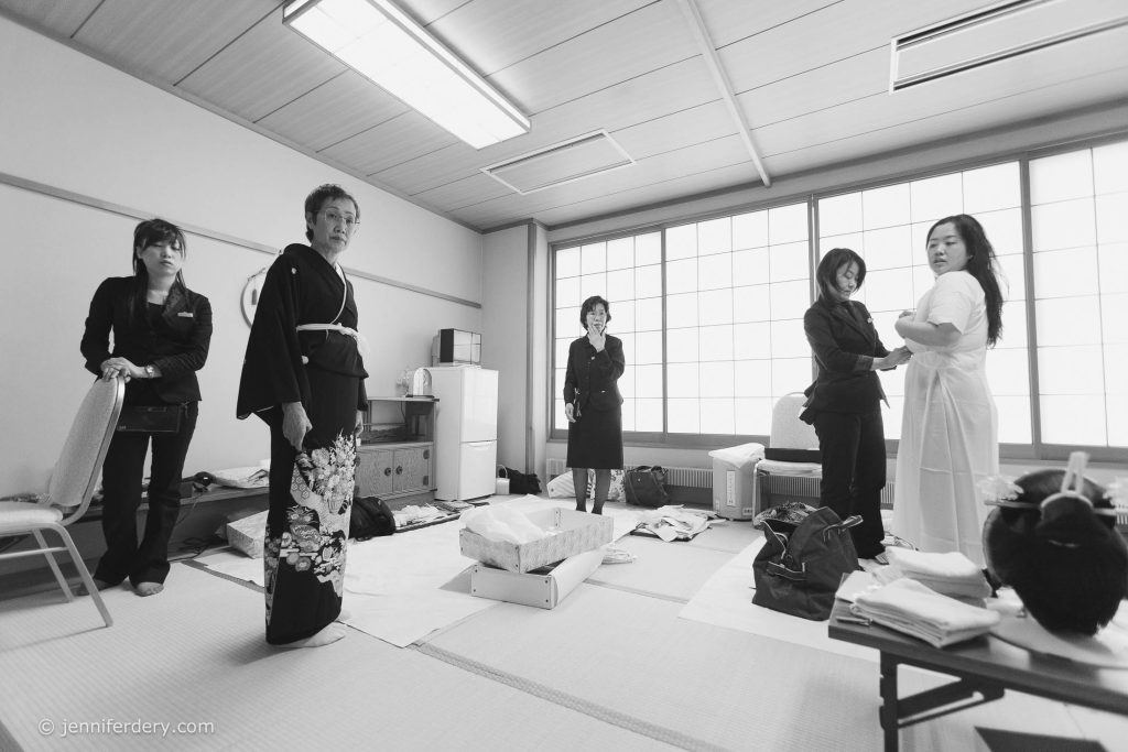 Five women stand in a tatami room with shoji screens, some in black attire and one in a white robe being assisted. Items and boxes are scattered around the floor, creating a sense of preparation or dressing.