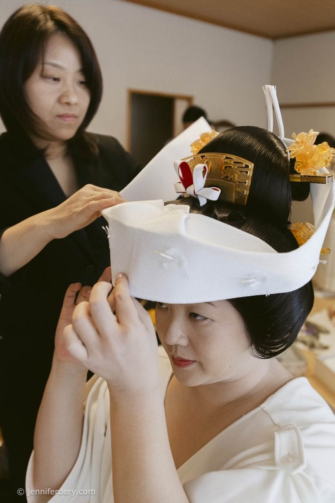 A woman helps a bride adjust her traditional Japanese wedding headpiece, known as a tsunokakushi, in a room. The bride wears white and has an elaborate hairstyle with decorative ornaments.