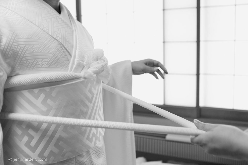 A person in a patterned kimono is being assisted by another person to tie a wide sash around their waist, inside a room with shoji screens. The scene is in black and white.