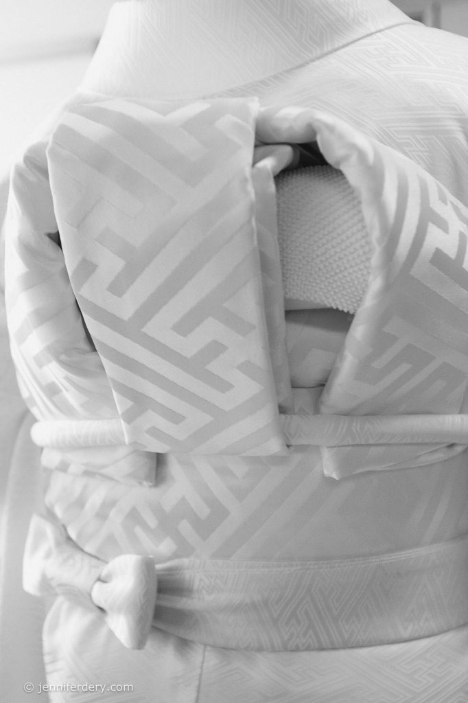 A close-up of the back of a person wearing a traditional Japanese kimono with a decorative obi bow. The fabric features a geometric pattern and the image is in black and white.