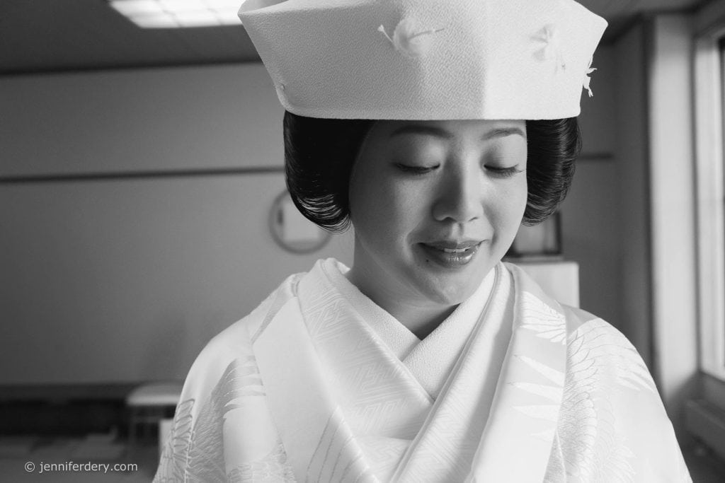 A woman in traditional Japanese bridal attire stands indoors, looking down with a serene expression. She wears a white kimono and a decorative hat called a tsunokakushi. The image is in black and white.