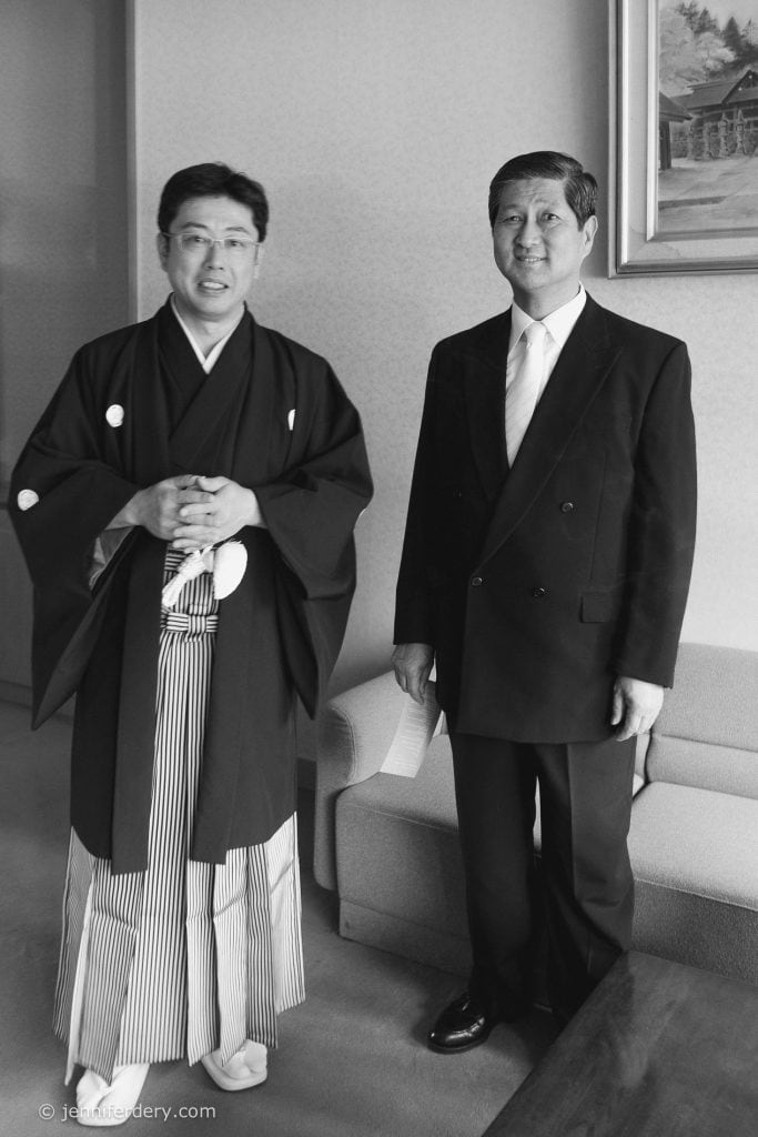 Two men stand indoors; one wears traditional Japanese attire with a kimono and hakama, while the other wears a black suit and tie. They are in a room with a sofa and framed artwork on the wall.