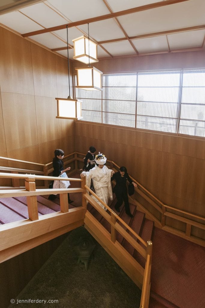 A bride in traditional Japanese wedding attire ascends a wooden staircase, assisted by three attendants dressed in black, in a bright, wood-paneled interior with large windows and hanging square lanterns.