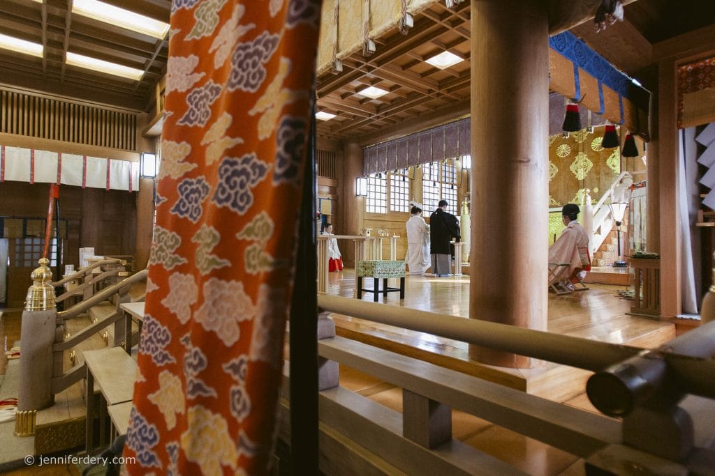 Inside a traditional Japanese shrine, people in white robes participate in a ceremony. Decorative fabrics and wooden architecture surround the scene, with natural light coming through the windows.
