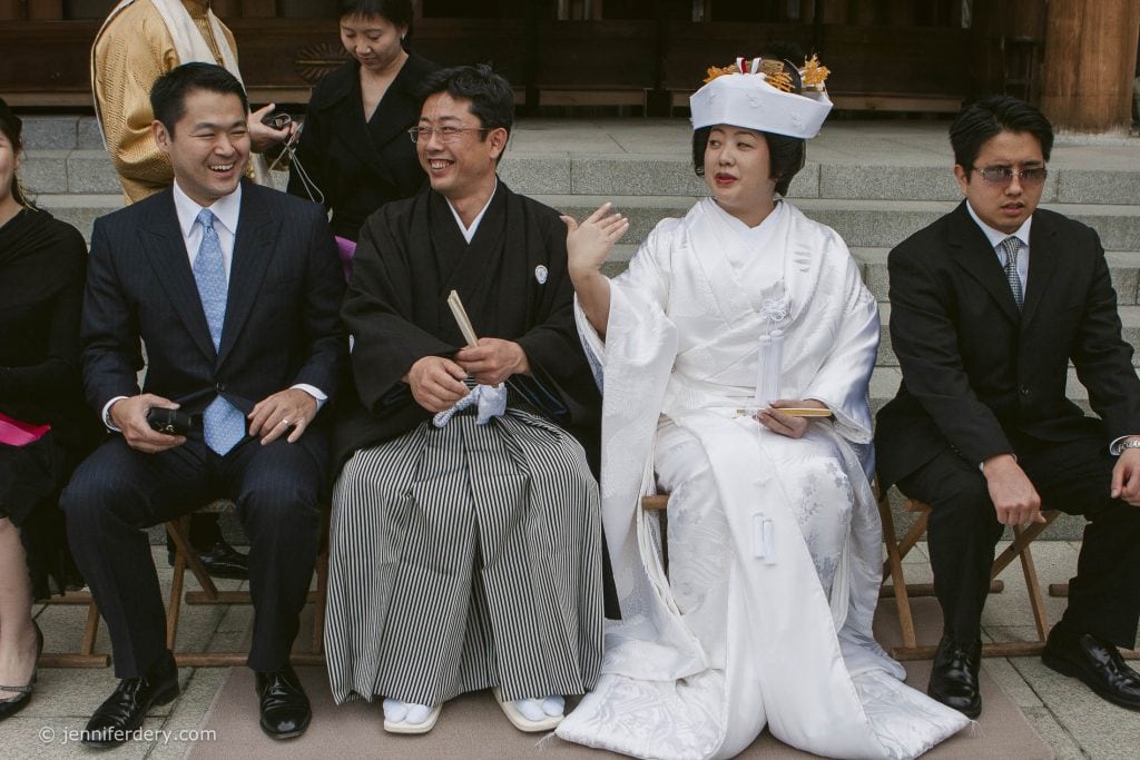 A group of people sits outside on wooden chairs. A bride in a traditional white Japanese wedding kimono and headdress gestures to a groom in black kimono. Others nearby are dressed in formal attire, smiling and chatting.