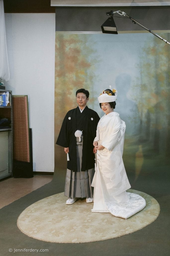 A couple poses in a photo studio wearing traditional Japanese wedding attire; the groom is in a black kimono and the bride in a white kimono with a decorative headpiece, standing on a circular rug with a nature backdrop.