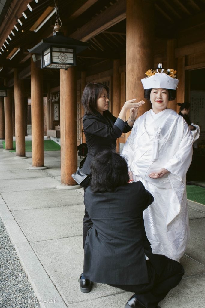 A bride in a traditional white Japanese wedding kimono and headpiece stands while two people in black adjust her attire and hair outside a wooden building with columns.